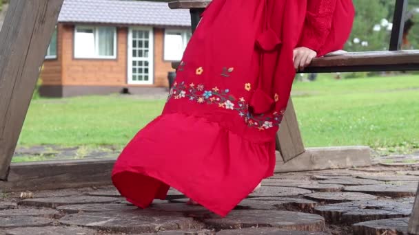 Fată într-o rochie etnică roșie stă pe un leagăn de lemn în parc — Videoclip de stoc