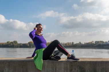 Koşu sporundaki genç ve yakışıklı koşucu kadın nehrin kenarında oturur ve güzel güneşli bir günde kardiyo antrenmanından sonra mola verir. 