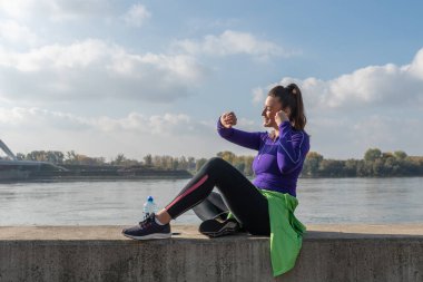 Koşu sporundaki genç ve yakışıklı koşucu kadın nehrin kenarında oturur ve güzel güneşli bir günde kardiyo antrenmanından sonra mola verir. 