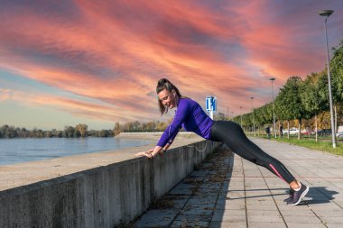 Genç, güçlü, atletik, aktif, sportif bir kadın. Açık hava egzersizi olarak nehir kenarında şınav çekiyor. Güzel bir gün batımı egzersizi yapıyor.