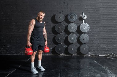 Genç, aktif, güçlü, terli, iri kaslı, spor salonunda duran ve elinde iki eski ağır su ısıtıcısı çanı tutan kaslı adam yoğun çapraz antrenmana hazırlanıyor.