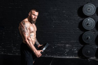 Genç, aktif, terli, güçlü, kaslı, elinde ağır halter plakası tutan kaslı adam spor salonundaki ağır çapraz omuz antrenmanına hazırlanıyor.