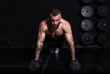 Genç, aktif, terli, formda, kaslı, kaslı bir adam spor salonunun dambıllarında şınav çekiyor.