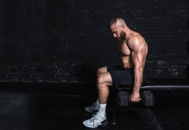 Genç, aktif, güçlü, terli, kaslı, iri kaslı bir adam bankta oturuyor ve spor salonundaki dambıllarla kas egzersizi yapıyor.