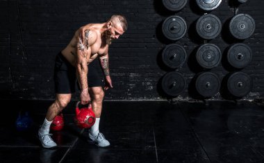 Genç, aktif, terli, güçlü, kaslı, kaslı, arka omuz kaslı, spor salonunda ağır bir su ısıtıcısı çanı ile antreman yapan bir adam.