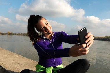 Sporda genç ve aktif bir bayan spor yaparken açık havada oturup, spor egzersizlerinden sonra akıllı telefonları dinleyerek ve antrenmanlara ara vererek selfie çekiyor.