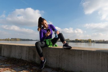 Genç, terli, aktif atletik kadın dışarıda koşma egzersizi yaptıktan sonra ara veriyor ve akıllı telefonundaki müziği değiştiriyor.