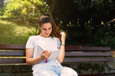 Genç mutlu kadın parkta bankta oturuyor ve güneşli günün tadını çıkarıyor. 