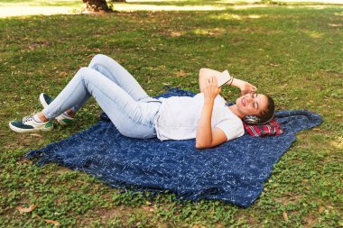 Genç, güzel, mutlu bir kadın kafasında kablosuz kulaklıkla parkta uzanıyor müzik dinliyor ve eğleniyor.