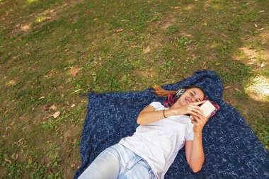 Genç, güzel, mutlu bir kadın kafasında kablosuz kulaklıkla parkta uzanıyor müzik dinliyor ve eğleniyor.