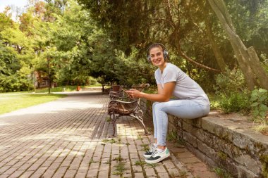 Genç, güzel, modern bir kadın. Parkta oturmuş, kablosuz kulaklıklarıyla akıllı telefonundan müzik dinliyor ve gülümsüyor.