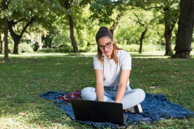 Genç ve güzel bir kız öğrenci, parkta battaniyenin üzerinde oturuyor ve internette dizüstü bilgisayarında bir sonraki sınav için belgeleri arıyor.