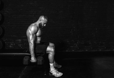 Genç, aktif, güçlü, terli, kaslı, iri kaslı bir adam bankta oturuyor ve spor salonunda dambıllarla antreman yapıyor.