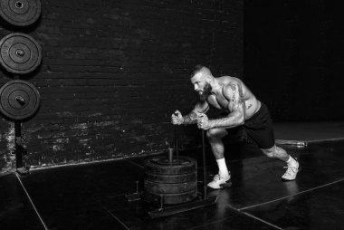 Genç, aktif, terli, iri kaslı, kaslı bir adam spor salonundaki ağır ağırlık plakalarını itiyor. Çapraz egzersizler yaparak gerçek insanları çalıştırıyor.