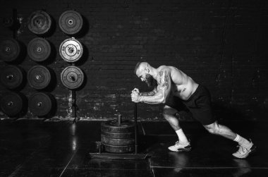 Genç, aktif, terli, iri kaslı, kaslı bir adam spor salonundaki ağır ağırlık plakalarını itiyor. Çapraz egzersizler yaparak gerçek insanları çalıştırıyor.