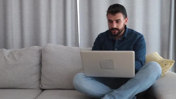 自宅のリビングルームに座っている若い創造的な幸せなフリーランスの男性と彼の会社のための新しいスタートアッププロジェクトのアイデアで残業ラップトップコンピュータを使用して 自宅のコンセプトで作業 — ストック動画