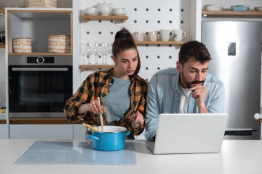 Genç çift mutfakta dizüstü bilgisayarda internet tarifini izlerken makarna ile yeni bir tabak pişiriyorlar ve kadın fırın tutuyor ve karıştırıyor. 