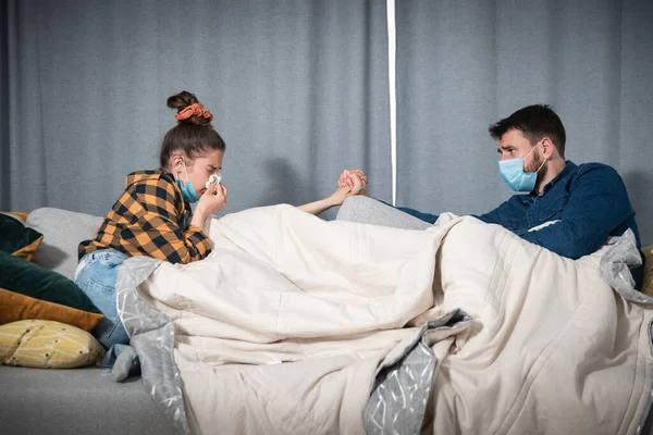 Young couple lies on the bed at a distance covered with a blanket with protective medical masks on their faces because they are sick with the coronavirus or covid-19 virus having health issues