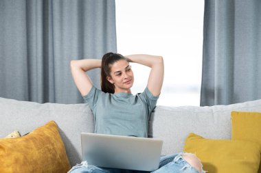 Genç ve mutlu serbest girişimci kadın evinde dizüstü bilgisayarla oturuyor ve gülümsüyor çünkü internetten bulduğu bir iş için çok önemli bir proje ödevini tamamladı.
