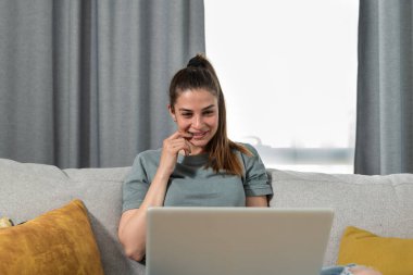 Genç ve mutlu serbest girişimci kadın evinde dizüstü bilgisayarla oturuyor ve gülümsüyor çünkü müşteri için çok önemli bir iş projesini ve internetten bulduğu bir işi tamamladı.