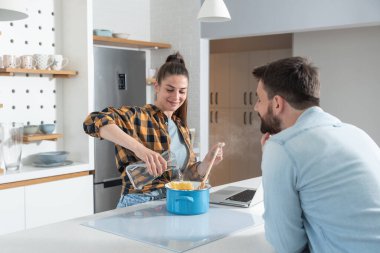 Mutlu genç çift akşam yemeği için mutfakta makarna pişirirken ve internetten yemek tarifleri okurken eğleniyor.