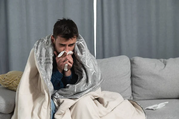 一名患有感冒 流感或过敏的年轻人坐在家里 裹着毛毯 用纸巾擦拭鼻子 而他却有强烈的头痛和健康意识 — 图库照片