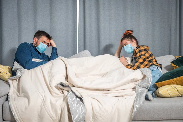 Young couple lies on the bed at a distance covered with a blanket with protective medical masks on their faces because they are sick with the coronavirus or covid-19 virus having health issues