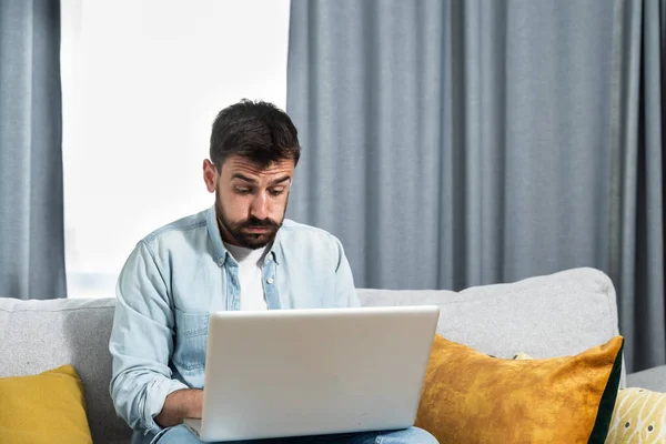 Young stressed freelancer business man working from home looking at his laptop computer screen reading negative charts and statistic after crisis and crash of the stock market