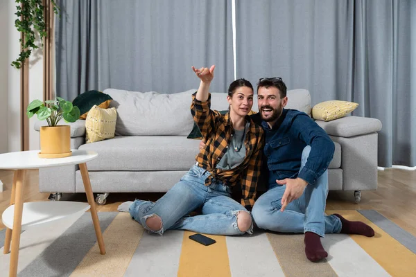 Young couple sitting on the floor of their home and watch love movie on television while woman enjoying and explaining to him the scene while he pretend to be interested with fake smile on his face