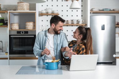 Genç mutlu çift mutfakta durup yemek pişiriyor. İnternette buldukları tarifleri kullanıyorlar.
