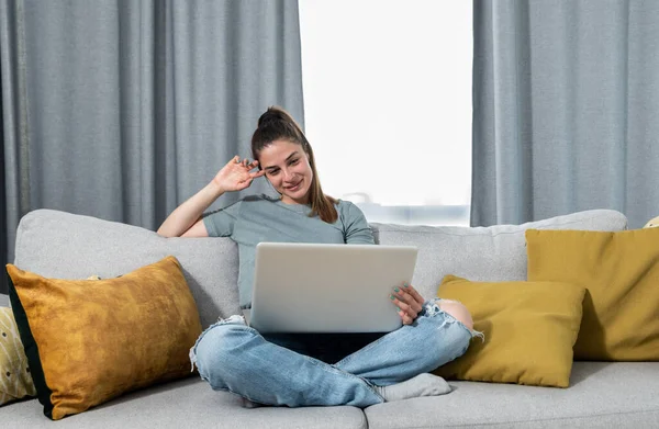 Young beautiful woman sitting on a sofa in apartment and watches her favorite series online on a laptop over the internet and enjoys her free day off for the weekend without worries and obligations