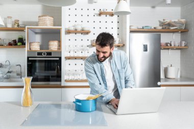 Genç mutlu adam mutfağında akşam yemeği için spagetti pişiriyor ve yemek bloguna koymak için bulduğu yeni tarifi laptopuna yazıyor.