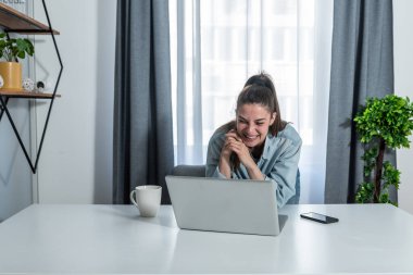 Genç, mutlu, serbest çalışan iş kadını ev ofisteki işine ara veriyor ve internetteki video konuşmasında iş arkadaşıyla sohbet ederek fincandan kahve içiyor. 