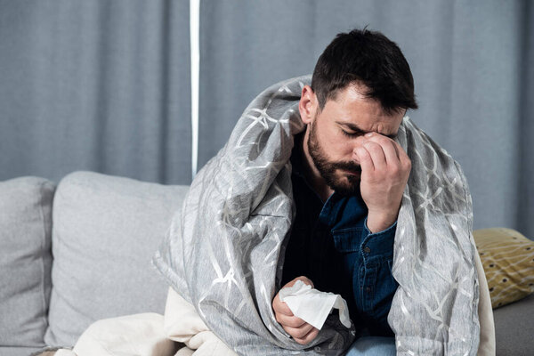 Молодой человек, страдающий обычной простудой и гриппом или аллергией, сидит дома, завернутый в одеяло и вытирает нос салфетками, пока у него сильная головная боль, концепция здравоохранения