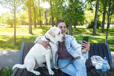 Evcil köpeğiyle oturan genç ve güzel bir kadın bankta altın renkli bir Labrador Retriever akıllı telefonuyla selfie çekmek istiyor. 