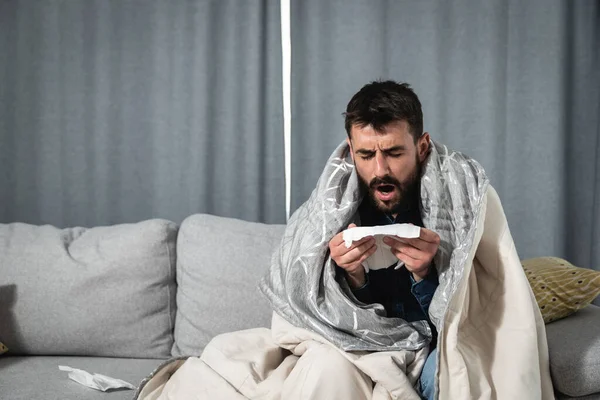 一名患有感冒 流感或过敏的年轻人坐在家里 裹着毛毯 用手帕擦鼻涕 而他却有强烈的头痛和健康意识 — 图库照片