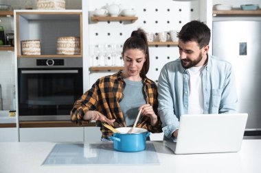 Genç mutlu çift mutfakta durup yemek pişiriyor. İnternette buldukları tarifleri kullanıyorlar.