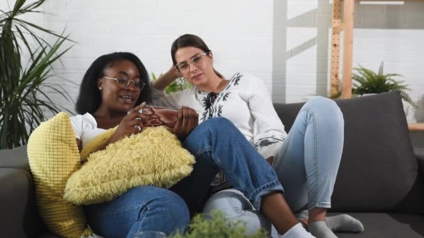 在同性恋关系中 一对年轻的非洲黑人和白人女性夫妻坐在沙发上 一边在智能手机屏幕上观看视频 一边享受他们的闲暇时光 — 图库视频影像
