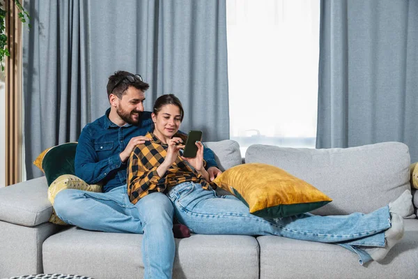 Young happy couple man and woman relaxing on the sofa in their home surfing the internet on the smartphone looking for funny videos and memes to have quality free time on weekend