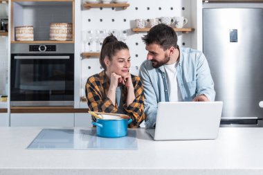Genç ve mutlu bir çift, yemek pişirmek için dizüstü bilgisayarlarında yemek tarifleriyle çevrimiçi kurs izliyor.