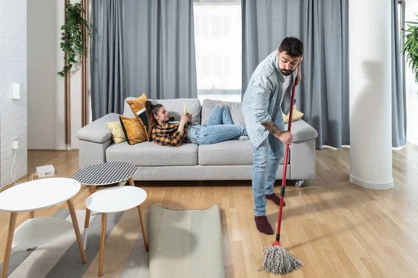自宅の若いカップル男は床を拭くと掃除するので 彼の女性は休憩し 彼女のスマートフォンでインターネットを閲覧し 彼はほこりや汚れから床をきれいにすることができますようにソファに横たわっていることができます — ストック写真