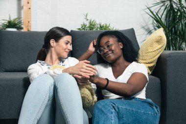 Genç ve güzel melez lezbiyen çift evlerinin zemininde oturup el ele tutuşurken ve gülümserken ve seçici bir şekilde odaklanırken ilişkilerinin tadını çıkarıyorlar.