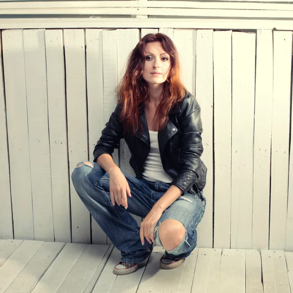 Beautiful young woman in jeans sitting against grunge wood wall — Stock Photo, Image