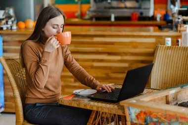 Genç ve güzel bir kız kahve içiyor ve kafede dizüstü bilgisayarda çalışıyor.