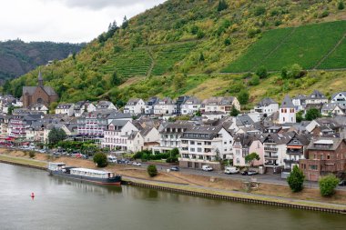 Moselle Nehri boyunca uzanan büyüleyici kasabanın panoramik manzarası ve Cochem, Almanya 'da bulutlu bir gökyüzünün altındaki sarp, teraslı üzüm bağları.