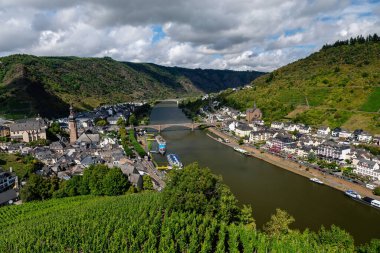 Moselle Nehri boyunca uzanan büyüleyici kasabanın panoramik manzarası ve Cochem, Almanya 'da bulutlu bir gökyüzünün altındaki sarp, teraslı üzüm bağları.