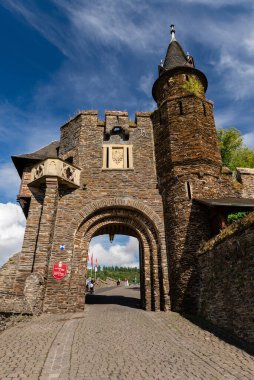 Cochem, Almanya - 9 Eylül 2025: Beyaz bulutlu dramatik mavi gökyüzü altında tarihi Reichsburg Cochem kalesinin heybetli taş kapı ve kule girişi, Almanya. Turistler ana kemer yolunda yürürken görülebilir.