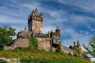 Orta Çağ ve Neo-Gotik mimarinin çarpıcı bir örneği olan Reichsburg Cochem Kalesi 'nin heybetli merkezi kulesi ve taş duvarları, Almanya' nın Cochem kentindeki dramatik mavi gökyüzüne karşı yeşil yamaçtan yükseliyor..