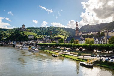 Moselle Nehri 'ndeki Cochem kasabasının geniş panoramik manzarası, tepedeki Reichsburg Cochem şatosu, St. Martin Kilisesi kulesi ve Almanya' nın güneşli sahil şeridi boyunca demirlemiş nehir tekneleri.
