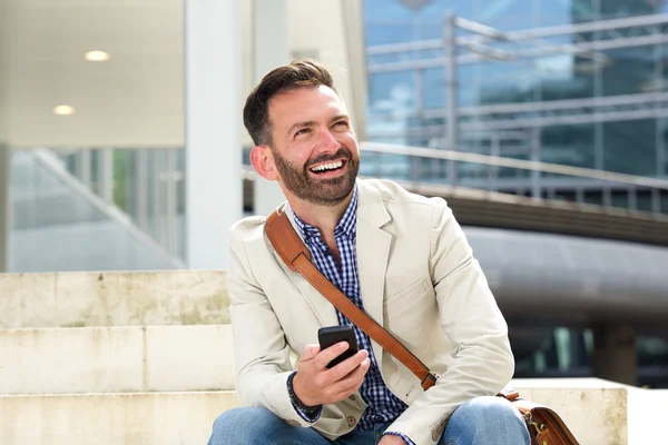 Śmiejąc się dojrzały mężczyzna siedzi na zewnątrz z cellphone — Zdjęcie stockowe